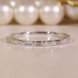 Peut inclure: Une bague en argent délicate avec une bande de pierres précieuses rectangulaires et transparentes. La bague est posée sur un fond doux et neutre, avec des orbes blancs flous en arrière-plan. La bague est de conception simple et élégante.