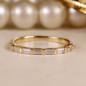 以下が含まれることがあります： 長方形の透明な宝石がバンドの周りにセットされたゴールドバンドリング。リングは、背景にぼやけた真珠がある明るい色の表面にあります。繊細なジュエリーです。