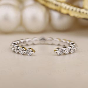 May include: A silver open ring featuring a line of clear, round gemstones. The ring has a gap in the middle and is set against a soft, neutral backdrop. The gemstones sparkle, reflecting light.