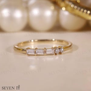 May include: A gold ring with a thin band, adorned with three rectangular-cut stones and two small round stones. The ring is presented on a neutral surface. The ring is a piece of jewellery.