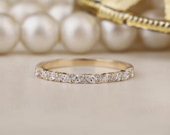 May include: A gold band ring with a row of alternating marquise and round-cut clear gemstones. The ring is set against a blurred background of pearls and gold jewelry.
