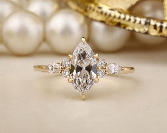 May include: A gold engagement ring with a large, marquise-cut diamond centre stone. The ring features smaller diamonds on either side of the centre stone. The band is a simple, polished gold. The ring is set against a soft, neutral background.