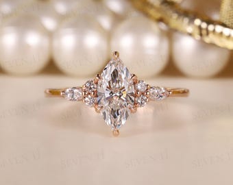 May include: A rose gold engagement ring with a marquise-cut central stone and smaller accent stones. The ring has a delicate band and a cluster of stones around the central diamond. The ring is set against a blurred background.