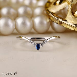 May include: A silver ring with a curved design, adorned with small, clear stones and a central, pear-shaped, deep blue gemstone. The ring's delicate details are set against a soft, neutral background, showcasing its elegance.
