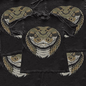 May include: A black short-sleeved t-shirt featuring a large, centred graphic of a snake head. The snake is depicted with detailed scales in shades of olive green, brown, and cream, with piercing yellow eyes and a focused, forward-facing expression.