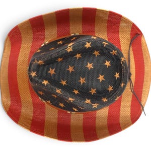 May include: Cowboy hat with a patriotic design. The hat features a black crown adorned with gold stars, and a brim with red and gold vertical stripes. A brown string is attached to the hat.