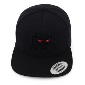 May include: Black snapback hat with a flat brim. The hat features an embroidered panther face with red eyes. A white and silver sticker is on the brim. The hat is made by Snapback.