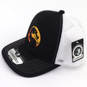 May include: Black and white trucker hat with a black and orange dragon logo on the front. The hat has a white mesh back and a black brim. The hat has a tag on the side that says "Cambridge" and "High Quality Premium Headwear".