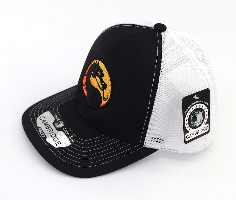 May include: Black and white trucker hat with a black and orange dragon logo on the front. The hat has a white mesh back and a black brim. The hat has a tag on the side that says "Cambridge" and "High Quality Premium Headwear".