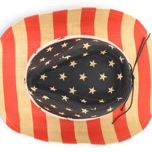 May include: A straw cowboy hat with a patriotic design. The brim features red and beige vertical stripes, while the interior crown is black with gold stars. A black string is attached for securing the hat.