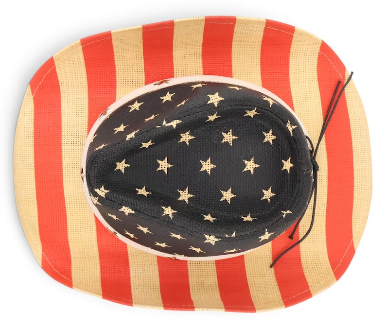 May include: A straw cowboy hat with a patriotic design. The brim features red and beige vertical stripes, while the interior crown is black with gold stars. A black string is attached for securing the hat.