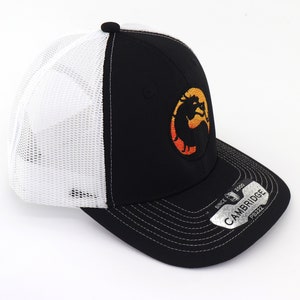 May include: Black and white trucker hat with a black and orange embroidered logo of a dragon. The hat has a white mesh back and a black brim with a silver tag that reads "CAMBRIDGE SINCE 2000 PB222".