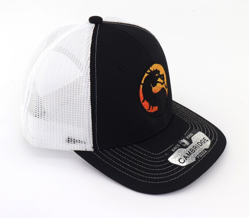 May include: Black and white trucker hat with a black and orange embroidered logo of a dragon. The hat has a white mesh back and a black brim with a silver tag that reads "CAMBRIDGE SINCE 2000 PB222".
