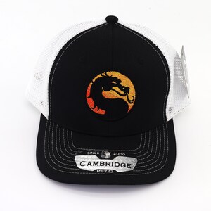 May include: Black and white trucker hat with a black dragon logo on a yellow and orange background. The hat has a white mesh back and a black brim. The hat has a label that says "SINCE 2000 CAMBRIDGE PB222".