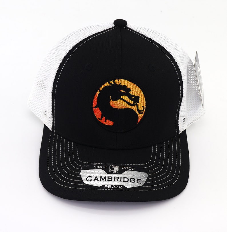 May include: Black and white trucker hat with a black dragon logo on a yellow and orange background. The hat has a white mesh back and a black brim. The hat has a label that says "SINCE 2000 CAMBRIDGE PB222".