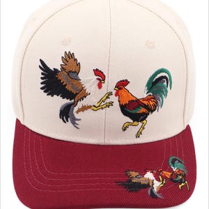 May include: A cream-colored baseball cap with a maroon brim and a maroon button on top. The front panel features an embroidered design of two roosters facing each other, while a smaller version of the same rooster embroidery is on the right side of the brim.