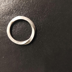 Puede incluir: Un anillo plateado con un diseño retorcido. El anillo es circular con un acabado liso y mate. El anillo está sobre un fondo oscuro.