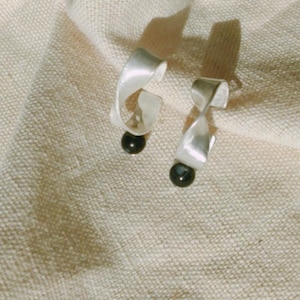 May include: A pair of silver-toned earrings with a ribbon-like design, each featuring a dark, round bead at the bottom. The earrings are set against a textured, off-white fabric background.