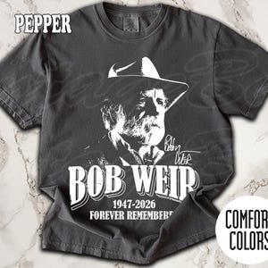 May include: A pepper-coloured t-shirt with a white graphic of Bob Weir in a hat, with his name, birth and death years, and the words "Forever Remembered." The shirt is from Comfort Colors.