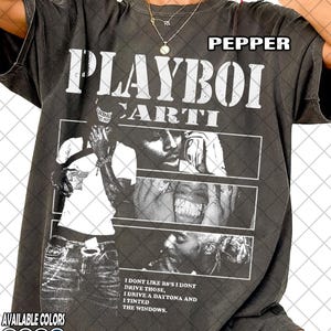 May include: Pepper-colored t-shirt featuring the text "PLAYBOI CARTI" in large white letters. The shirt includes a graphic of the artist and the text "I DONT LIKE R8'S I DONT DRIVE THOSE, I DRIVE A DAYTONA AND I TINTED THE WINDOWS." Available in multiple colors.