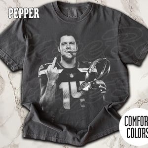 May include: Pepper-colored Comfort Colors t-shirt featuring a black and white image of a football player holding a trophy and cigar, with the number 14 on the jersey. The player is giving the middle finger. The shirt has the Comfort Colors logo.