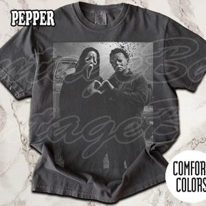 May include: Dark gray t-shirt with a black and white graphic of two horror movie characters forming a heart with their hands. The shirt has the word "PEPPER" printed at the top and "COMFORT COLORS" at the bottom.