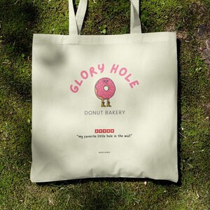 May include: Cream-colored tote bag with the words "GLORY HOLE" in pink above a pink donut illustration. The donut has a brown center and is wearing brown boots. Below the donut is "DONUT BAKERY". The bag also has the text "my favorite little hole in the wall".