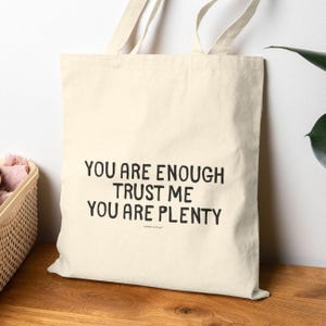 Puede incluir: Una bolsa de tela beige con texto negro que dice "You are enough trust me you are plenty".