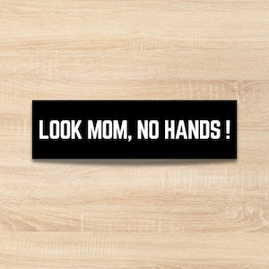 May include: A black rectangular sign with the phrase "LOOK MOM, NO HANDS!" in bold white letters. The sign is placed on a light brown wooden surface.