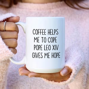 May include: White ceramic coffee mug with the text "COFFEE HELPS ME TO COPE POPE LEO XIV GIVES ME HOPE" in black lettering. The mug has a curved handle and is held by a person wearing a pink sweater.