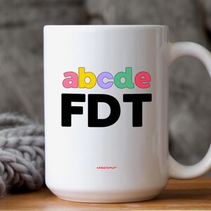 Puede incluir: Taza de café de cerámica blanca con un diseño negro y colorido. El diseño presenta las letras "abcde" en colores arcoíris y la palabra "FDT" en negro.