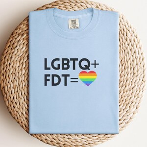 Puede incluir: Una camiseta azul claro con el texto "LGBTQ+ FDT =" y un corazón arcoíris. La camiseta está doblada y colocada sobre una superficie circular tejida. La camiseta tiene cuello redondo y mangas cortas.
