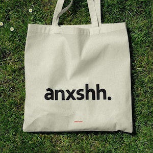 Puede incluir: Un bolso tote beige con la palabra "anxshh." impresa en negro. La frase "added humor" está impresa en rojo debajo. El bolso, con dos asas, está sobre hierba verde. Diseño sencillo y humorístico.