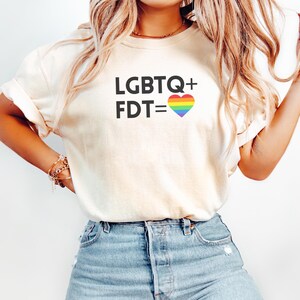 Op de afbeelding: Crèmekleurig T-shirt met de tekst "LGBTQ+ FDT=" en een regenboog hartafbeelding. Het shirt is gemaakt van een zacht materiaal en heeft korte mouwen. Het ontwerp is gecentreerd op de voorkant van het shirt.