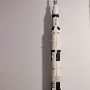 Può includere: Un modello di razzo bianco e nero con la scritta "USA" in rosso. Il razzo ha più stadi e alette, ed è una replica del razzo Saturn V. Il modello include bandiere americane ed è esposto contro un muro bianco.
