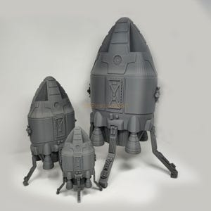 May include: Three gray, 3D printed, model spaceships of varying sizes. The largest spaceship has a rounded top and three legs. The middle spaceship has a rounded top and three legs. The smallest spaceship has a rounded top and three legs.