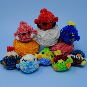 Pode incluir: Um grupo de brinquedos de pássaros de crochê coloridos, alguns sentados em bolas de lã. Os pássaros são feitos de lã em tons de vermelho, rosa, azul, verde e marrom.