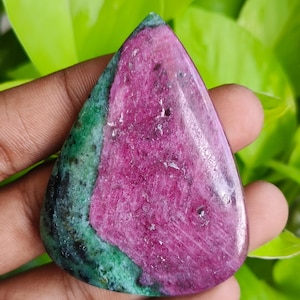 Könnte beinhalten: Ein polierter Edelstein in Tropfenform. Der Stein hat eine leuchtend magenta Mitte, die von einem grünen und schwarzen Band umrandet ist. Der Edelstein wird in einer Hand gehalten, mit grünem Laub im Hintergrund.