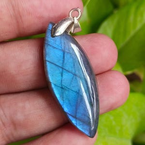 Puede incluir: Un colgante de labradorita pulida en forma de hoja que presenta un destello iridiscente azul vibrante en su superficie. La piedra preciosa está montada en un sencillo engaste de metal plateado en la parte superior, con una forma curva.