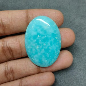 以下が含まれることがあります： 鮮やかなターコイズ色の、磨かれた楕円形の宝石。滑らかで光沢のある表面に、わずかに白い縞模様があり、灰色の背景に手で持たれています。宝石の長さは約3.8cmです。