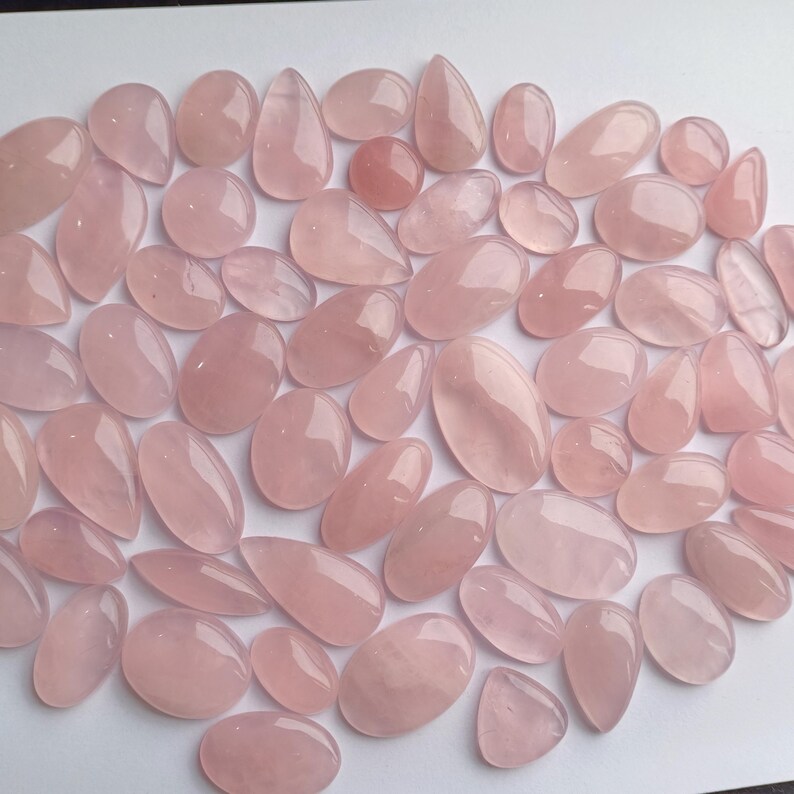 May include: A collection of pink rose quartz cabochons, a type of gemstone, in various sizes and shapes. The cabochons are smooth and polished, with a translucent appearance.