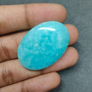 以下が含まれることがあります： 鮮やかなターコイズ色の、磨かれた楕円形の宝石。滑らかで光沢のある表面に、わずかに白いインクルージョンがあり、灰色の背景の中で手に持たれています。宝石の長さは約5cmです。