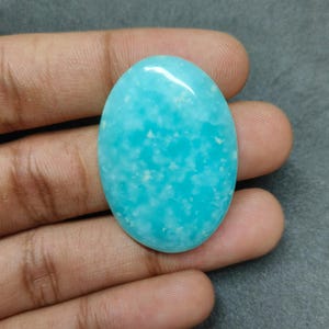 以下が含まれることがあります： 鮮やかなターコイズ色の、楕円形に研磨された宝石。滑らかで光沢のある表面を持ち、淡い色のインクルージョンが散りばめられた効果を生み出しています。宝石は手で持たれています。