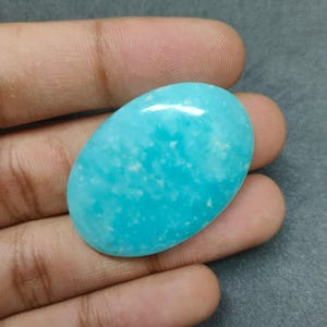 以下が含まれることがあります： 鮮やかなターコイズ色の、楕円形に研磨された宝石。 石は明るく鮮やかな色で、より明るいインクルージョンがあります。 宝石は手で持たれており、そのサイズと質感が示されています。 長さ約4cm。