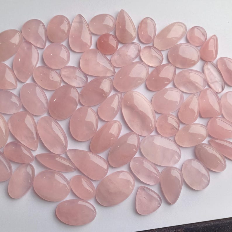 May include: A collection of pink rose quartz cabochons, a type of gemstone, in various shapes and sizes. The stones are smooth and polished, with a translucent appearance.