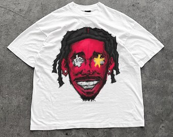 May include: White t-shirt featuring a red face graphic with black hair and a smiling expression. The eyes have a star and a symbol design. The graphic tee is a casual wear item.
