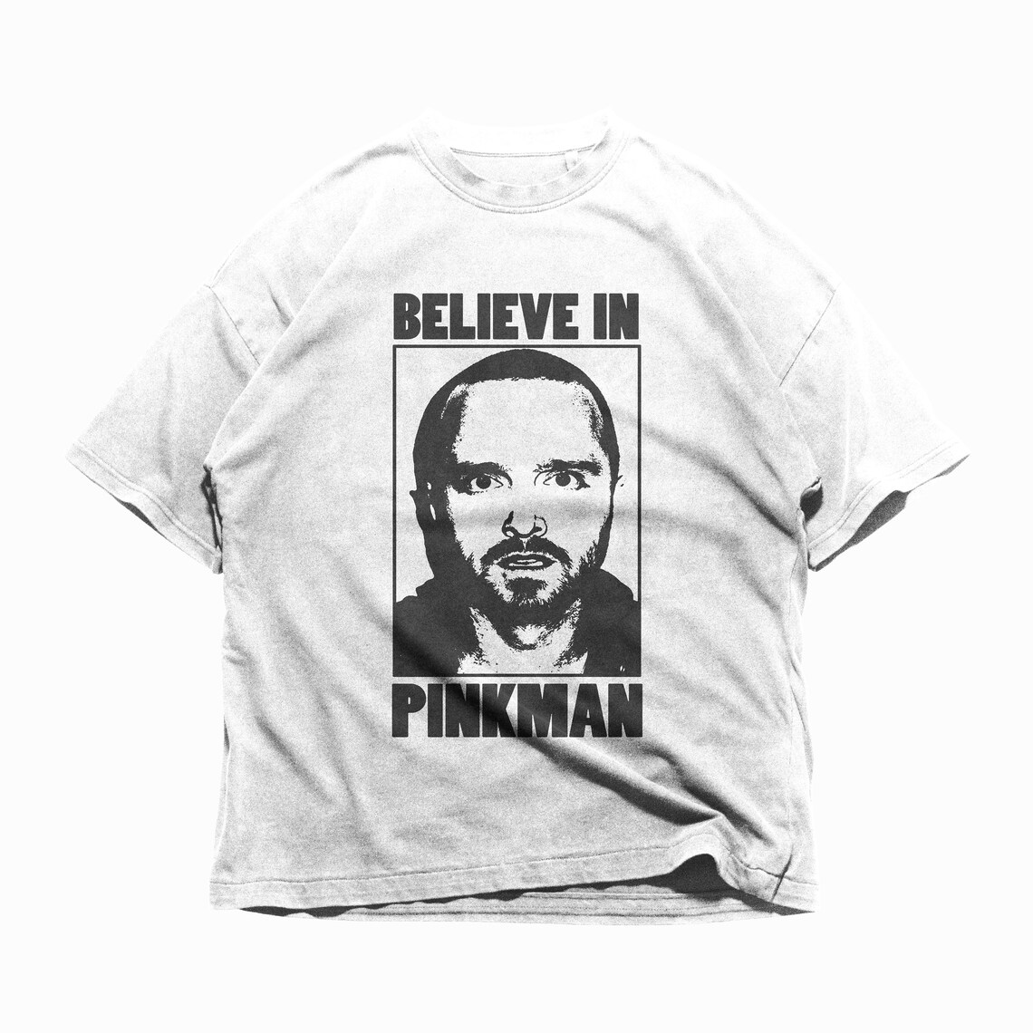 Op de afbeelding: Wit t-shirt met een zwart-witte afbeelding van een mannengezicht in een rechthoekige lijst. De tekst "BELIEVE IN PINKMAN" staat boven en onder de afbeelding. Het shirt heeft korte mouwen en een ronde hals.