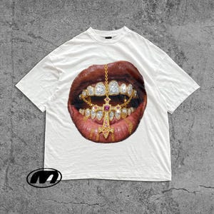 Puede incluir: Camiseta blanca con un gráfico de una boca con dientes de oro y un colgante de cruz. Los labios son de color rojo-marrón oscuro, y los dientes están adornados con diamantes. El colgante tiene una gema rosa en el centro.
