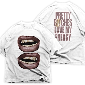 Könnte beinhalten: Weißes T-Shirt mit einem Motiv von zwei Lippenpaaren mit goldenen Zähnen auf der Vorderseite und dem Text "PRETTY BITCHES LOVE MY ENERGY" auf der Rückseite.