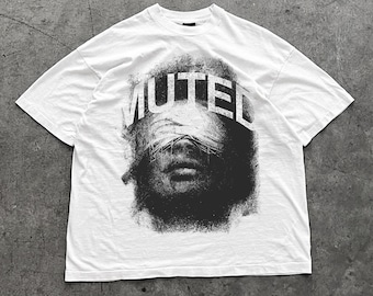 Op de afbeelding: Wit T-shirt met een zwart-witte afbeelding van een gezicht met het woord "MUTED" over de ogen. De afbeelding heeft een verweerde, getextureerde uitstraling, wat een krachtige visuele impact creëert. Het shirt heeft een klassieke ronde hals.